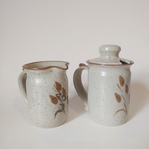 May include: A set of two ceramic creamer and sugar containers with brown floral designs on a white speckled background.