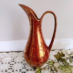 May include: A tall, slender pitcher with a glossy, metallic copper and bronze finish featuring a distinct mottled, circular pattern. The vessel has a curved handle, a wide, rounded base that tapers towards the top, and a prominent, elongated pouring spout.