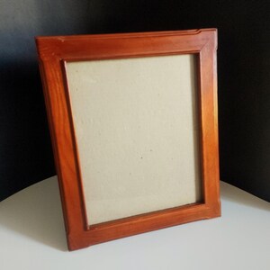 May include: A rectangular picture frame with a smooth, medium-toned reddish-brown wooden finish. The frame features a simple, clean-lined design with mitred corners and a plain, light beige mat board insert centred within the frame opening.