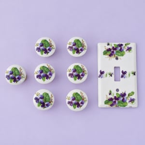 May include: A set of six round white ceramic cabinet knobs and one rectangular white ceramic light switch plate, each decorated with a painted pattern of purple violet flowers and green leaves against a clean white background.