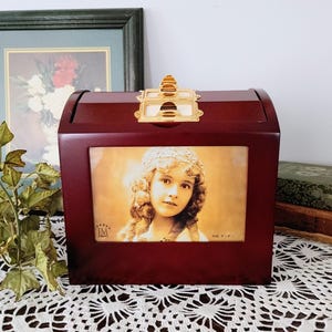 Peut inclure: Une boîte à souvenirs en bois couleur cerise foncée avec un loquet en métal doré sur le dessus. L'avant présente un cadre photo rectangulaire contenant le portrait d'une jeune personne aux cheveux bouclés. Le texte "JORDAN JM MARK" est visible.