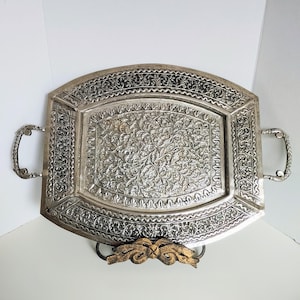 May include: Silver metal tray with detailed floral and scroll patterns. The rectangular tray has curved edges and handles. The tray is supported by a decorative stand with a gold-coloured accent. The tray's dimensions are approximately 40 cm x 30 cm.