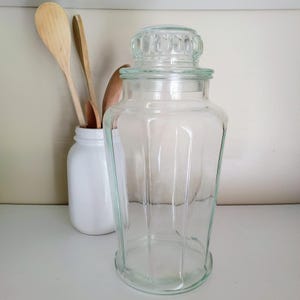 May include: A clear glass jar with a lid, next to a white jar holding wooden spoons. The jar has a decorative lid and a wide base. The spoons are light brown. The background is a neutral color.