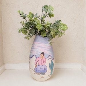 May include: A decorative ceramic vase with a hand-painted design featuring a woman, mountains, and a cactus. The vase is filled with green leafy stems. The color palette includes pink, blue, and beige.