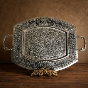 May include: An oval silver-toned metal serving tray featuring intricate embossed floral and vine patterns across the entire surface. The tray includes two decorative side handles and a raised border, supported by a gold-toned ornate base.