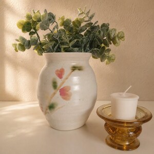 May include: A white ceramic vase with a floral design, holding green leafy branches, sits next to a small amber glass candle holder with a white candle. The vase has a hand-painted floral pattern in pink and green.