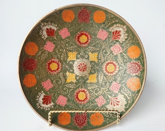 May include: Decorative round metal plate with a green background and intricate floral design in orange, red, pink, and yellow. The plate has a gold rim and is displayed on a gold stand.