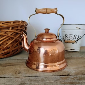May include: A polished copper tea kettle with a rounded body, a curved spout, and a hinged handle featuring a light-coloured wooden grip. The kettle has a matching copper lid with a small knob and is positioned in front of a wicker basket.