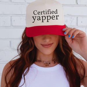 May include: A cream-coloured baseball cap with a bright red brim and a small red button on top. The front of the cap features the text "Certified yapper" printed in a bold, black, serif font. The cap has a structured crown and a curved visor.