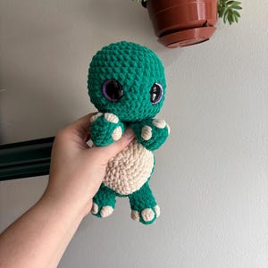 Pode incluir: Um brinquedo de pelúcia de tartaruga verde de crochê com detalhes brancos e grandes olhos roxos.