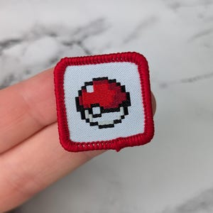 May include: Square embroidered patch featuring a pixelated red and white Pokeball design on a light blue background. The patch has a red border. The patch is held by a person's hand.