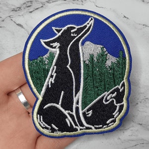 May include: Embroidered patch featuring a black wolf howling at the moon, with a blue sky and green trees in the background.