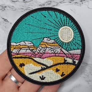 May include: Embroidered patch depicting a colourful mountain landscape with a sun and rays. The mountains are pink, yellow and white, and the sky is turquoise. The sun is white with a yellow outline.