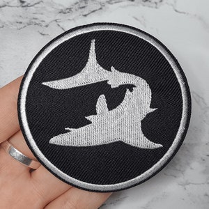 May include: Black and white embroidered patch featuring two stylised sharks facing each other. The patch is circular and has a white border.