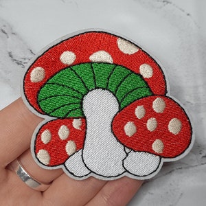 Könnte beinhalten: Gestickter Aufnäher mit drei roten und weißen Pilzen mit grünen Stielen. Die Pilze sind übereinander gestapelt.