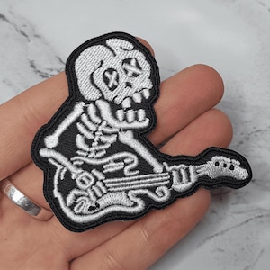 Op de afbeelding: Wit skelet dat een zilveren elektrische gitaar bespeelt met zwarte details. Het skelet heeft een schedelkop met X voor ogen. De patch is op een zwarte achtergrond.