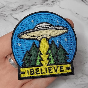 May include: Embroidered patch depicting a UFO above a forest, with the text "I BELIEVE". The patch has a blue sky with stars, a yellow light beam, and green trees. The UFO is beige.