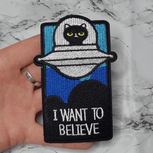 Op de afbeelding: Zwarte kat in een UFO met de tekst "Ik wil geloven" op een blauw en zwart geborduurd patch.