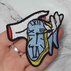 May include: Embroidered patch depicting a melting clock design in blue, yellow, and black, held by white hands with black outlines. The clock face shows Roman numerals. The patch has a black border and a red accent.