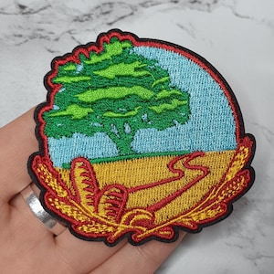Op de afbeelding: Geborduurde patch met een groene boom tegen een blauwe lucht en een geel veld. Een rode weg slingert door het veld. De patch is omlijst door een rood en goudkleurig ontwerp met een zwarte omtrek.