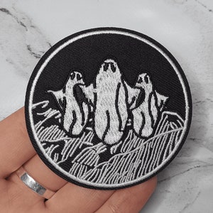 May include: Black and white embroidered patch featuring three ghosts in a row, with a white background and black outlines. The ghosts are in a spooky, Halloween-themed design.
