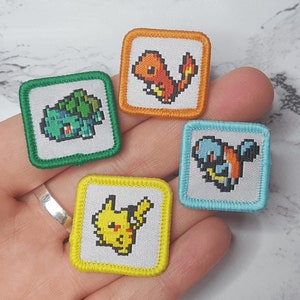 May include: Four embroidered iron-on patches featuring pixelated Pokemon characters. The patches are green, yellow, blue, and orange. The characters are Bulbasaur, Pikachu, Squirtle, and Charmander.
