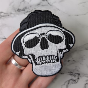 Può includere: Toppa ricamata con un teschio bianco che indossa un cappello nero. Il teschio ha orbite oculari dettagliate, denti e una mascella definita. La toppa è delineata in bianco e nero, creando un contrasto sorprendente. La toppa è probabilmente per abbigliamento o accessori.