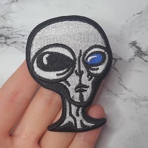 May include: Embroidered iron-on patch of a grey alien with one blue eye and one black eye.