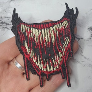 May include: A black and red embroidered patch featuring a large, toothy, open mouth dripping with red and black liquid. The mouth is outlined in white.