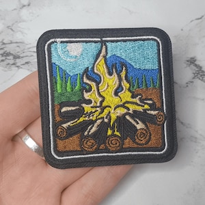 Peut inclure: Écusson brodé représentant une scène de feu de camp. Le motif comprend une flamme jaune et orange, des bûches marron, des arbres verts, des montagnes bleues et un ciel bleu clair avec une lune blanche. L'écusson a une bordure noire.