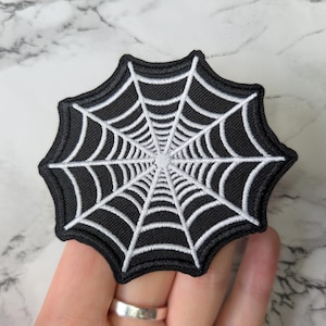 Pode incluir: Patch bordado de teia de aranha em preto e branco. O patch apresenta um design detalhado de teia de aranha branca sobre um fundo preto. O patch é circular com uma borda ondulada. Ideal para o Halloween ou projetos góticos.