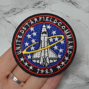 May include: Embroidered patch with the text "UNITED STARFIELD COMMAND" and "1983". The patch features a white space shuttle design on a blue background with white stars and a yellow orbital path. The patch has a black border with red and white accents.