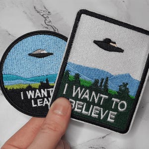 Op de afbeelding: Twee geborduurde patches met UFO-ontwerpen. Eén is cirkelvormig met een blauwe lucht en de tekst "I WANT LEAVE". De andere is rechthoekig met een landschap en de tekst "I WANT TO BELIEVE". Beide patches hebben een zwarte rand.