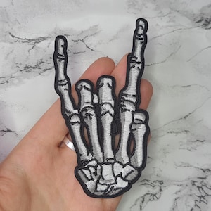 May include: A white skeleton hand making the rock and roll hand gesture. The hand is embroidered on a black fabric patch.