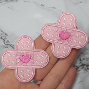 May include: Two pink felt iron-on patches shaped like bandages with a pink heart in the center. The patches have white stitching around the edges and white dots.