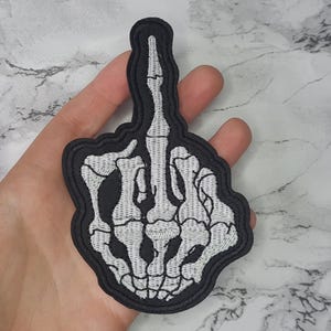 May include: A black and white embroidered patch of a skeleton hand giving the middle finger.