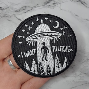 Könnte beinhalten: Schwarz-weiß gestickter Aufnäher mit einem UFO, einer Person, die darauf zugeht, und dem Text "I WANT TO LEAVE".