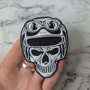 May include: Embroidered patch depicting a skull wearing a pilot's helmet and goggles. The patch is black and white, with detailed stitching outlining the skull's features and the helmet's design. The patch is held in a hand.