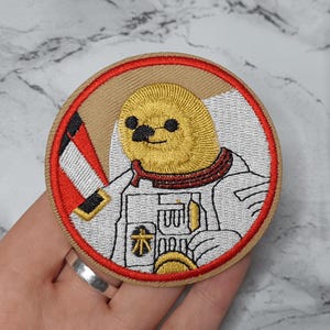 May include: A round embroidered patch with a beige background and a red border. The patch features a cartoon astronaut wearing a white spacesuit with a red collar. The astronaut has a yellow head with black eyes and a black moustache. The astronaut is holding a small object in its hand.