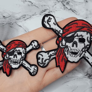 May include: Two embroidered iron-on patches featuring a skull and crossbones with a red bandana. The skull has a black eye patch and a toothy grin.