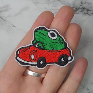 May include: Green frog embroidered patch riding in a red car. The car has white wheels and black tires. The frog is wearing a white hat.