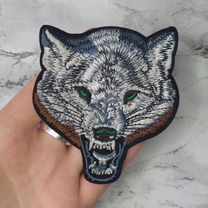 May include: Embroidered iron-on patch of a white wolf's head with green eyes, snarling with its mouth open, against a brown background.