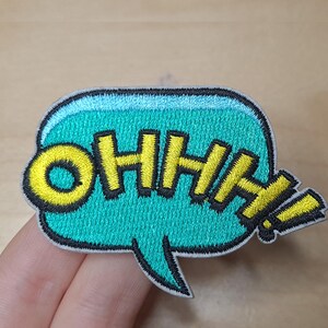 May include: A turquoise blue speech bubble with a yellow embroidered "OHHH!" inside. The speech bubble has a black outline and a white border.