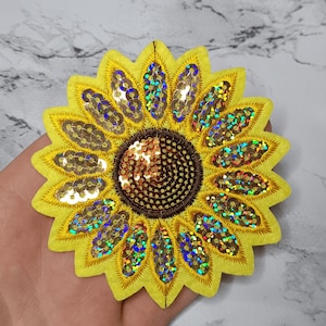 Puede incluir: Un parche de girasol de fieltro amarillo con lentejuelas iridiscentes. El centro del girasol es marrón y también está cubierto de lentejuelas.