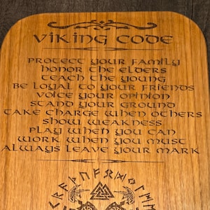 Könnte beinhalten: Eine Holztafel mit dem Wikinger-Kodex eingraviert. Der Kodex enthält Sätze wie "Schütze deine Familie", "Ehre die Ältesten" und "Hinterlasse immer deine Spuren". Die Tafel zeigt auch ein Wikingersymbol mit Runen.