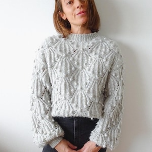 May include: A grey knitted jumper with a textured pattern and bobbles. The jumper has a round neckline and long sleeves.