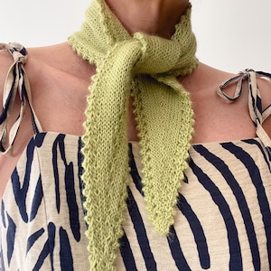 Può includere: Una sciarpa di maglia verde chiaro con un bordo a festoni. La sciarpa è annodata intorno al collo in un nodo morbido.