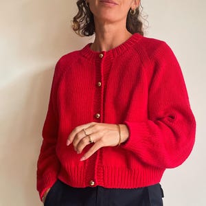 May include: A bright red cardigan sweater with gold-colored buttons. The sweater has a ribbed texture and a round neckline. The sleeves are long and the fit is cropped. The person is wearing dark blue pants.