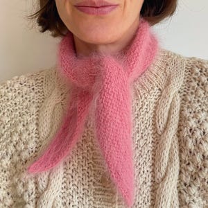 May include: A pink fluffy scarf tied around the neck of a person wearing a cream cable knit jumper.