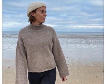 Könnte beinhalten: Eine Frau trägt einen braunen Rollkragenpullover mit einer weißen Mütze am Strand.